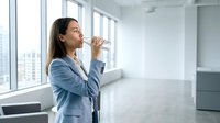 Professional Asian businesswoman drinking water in modern office space with city view windows for workplace wellness and hydration concepts.