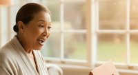 Mature woman of South Asian descent with dark hair pulled back, wearing a beige cardigan, smiling while holding a tablet device indoors near bright windows. Suitable for business, technology, and lifestyle content.