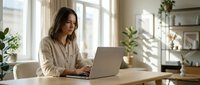 Young professional woman working on laptop at home office desk with natural lighting and plants creating productive workspace atmosphere.