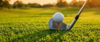 Golf club and ball on green grass course during golden hour sunset creating warm atmospheric lighting for sports and recreation activities.
