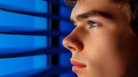Young Caucasian man looking through window blinds in dramatic blue lighting, profile view showing contemplative expression and strong facial features. Concept of hope and future.
