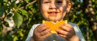 Young child with fair skin holding a freshly picked orange outdoors among green foliage. Ideal for agriculture, healthy eating, organic farming, and family lifestyle content.