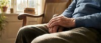 Senior man sitting peacefully in comfortable chair by sunny window, hands resting on lap in cozy home interior with warm natural lighting and plants.