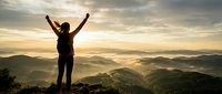 Young woman hiker celebrating success with raised arms on mountain summit at golden sunrise, overlooking misty valley landscape for motivation concepts.
