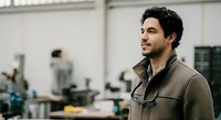 Young man with dark hair and facial hair, South Asian ethnicity, wearing brown work jacket in industrial workshop setting with machinery and tools in background.