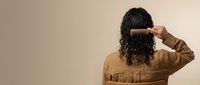 Woman with curly dark hair combing with wooden brush, rear view against neutral beige background for hair care and beauty routine concepts.