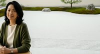 Middle-aged woman with dark hair and South Asian ethnicity wearing olive cardigan over white top, smiling at camera in serene zen garden with raked gravel patterns.