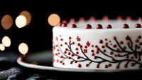 White frosted cake decorated with delicate red berry branch pattern on sides, bokeh lights in dark background, elegant holiday dessert presentation.