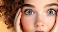 Close-up portrait of young woman with curly hair, amber eyes, and natural makeup expressing surprise with hands on face against yellow background.