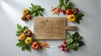 Wooden cutting board surrounded by fresh organic vegetables including tomatoes, carrots, radishes and herbs on white background for cooking preparation.