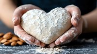 Heart shaped raw dough in female hands with flour on dark background and almonds. Close up view of homemade baking preparation process.