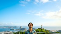 Young man with dark hair and medium skin tone wearing casual blue shirt, smiling while looking upward against scenic coastal landscape with mountains, lighthouse, and blue sky for travel and lifestyle content.