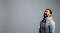 Young man with dark hair and beard, Middle Eastern ethnicity, wearing gray hoodie, smiling and looking upward against neutral background for lifestyle and portrait use.