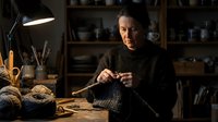 Middle-aged man with gray hair and facial hair, wearing dark turtleneck sweater, working on traditional rope or fiber craft at wooden table in studio workshop with shelves and task lighting.