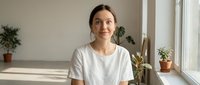 Young Asian woman in white t-shirt smiling at camera in bright modern apartment with houseplants by window, natural lighting for lifestyle content.
