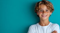 Happy young boy with curly blonde hair smiling confidently with arms crossed against turquoise background for youth lifestyle and education concepts.