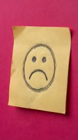 Yellow sticky note with hand-drawn sad face emoji on pink textured wall expressing negative emotions and disappointment for office communication.