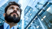 Young bearded businessman in blue suit looking up at modern glass office buildings with determined expression, urban corporate success concept.