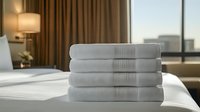 Fresh white folded towels stacked neatly on hotel bed with city view through window, warm interior lighting creates luxurious hospitality atmosphere.