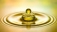 Golden oil drop splashing into liquid surface, creating crown ripples, macro shot with warm yellow tones for food, cosmetic, or wellness concepts.