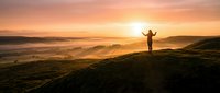 Person with arms raised standing on mountain ridge at sunrise overlooking misty valley landscape, celebrating achievement and freedom outdoors.