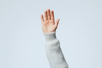 Raised hand in white knitted sweater sleeve against light background showing stop, vote, or participation gesture. Minimalist composition for social concepts.