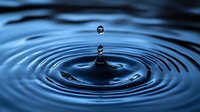 Water drop falling into calm blue water creating concentric ripple circles with secondary droplet suspended above, captured in macro photography.
