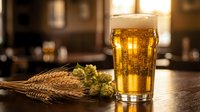 Fresh golden beer in glass with wheat stalks and hops on wooden table in warm pub setting. Craft brewing ingredients and alcoholic beverage concept.