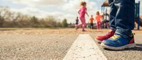 Young child wearing blue and red sneakers standing on asphalt road line with blurred group of children playing in background outdoors during daytime.