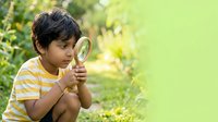 Young boy with dark hair and medium skin tone, wearing a yellow and white striped shirt, holding a magnifying glass while exploring outdoors in a garden setting. Suitable for educational materials, children's content, and discovery themes.