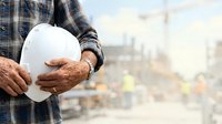 Worker in plaid shirt holding white safety helmet on construction site with blurred industrial background and equipment.