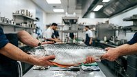 Young adult male with light skin tone holding a fresh salmon fish in a commercial kitchen environment with colleagues working in the background. Suitable for culinary, food industry, restaurant, and seafood business content.