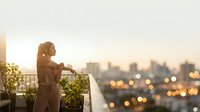 Woman in brown coat standing on urban rooftop balcony at sunset, gazing at blurred city skyline lights below, contemplative mood for lifestyle and aspirational concepts.
