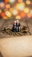 Miniature elderly couple figurines sitting on golden coins with treasure chests on wooden surface with warm bokeh lights background for retirement planning concept.