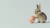 Young brown and white bunny sitting next to colorful Easter egg decorated with floral pattern on light neutral background. Minimal spring holiday composition.