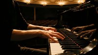 Close-up of male hands playing grand piano on stage in elegant concert hall with warm golden lights in background.
