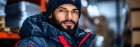 Young Hispanic male warehouse worker in winter jacket and beanie smiling confidently in industrial storage facility with blurred background.
