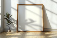 Empty golden frame leaning against white wall with window shadow and potted plant, minimalist interior design setup on wooden floor.