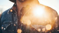 Young African American man in denim jacket silhouetted against bright sunset with bokeh light effects and falling snow creating dreamy winter atmosphere.