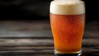 Cold glass of amber craft beer with thick white foam head and condensation droplets on dark wooden table, dramatic side lighting creates mood.