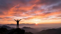 Silhouette of person with raised arms celebrating success on mountain peak at sunrise with dramatic orange and pink sky over layered mountain ranges.
