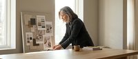 Senior woman with gray hair, wearing black blazer, working at wooden desk in bright modern office space, reviewing design samples and mood board on wall, professional interior designer or architect at work.