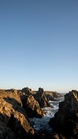 Dramatic rocky coastline with jagged cliffs and ocean waves under clear blue sky during golden hour light for travel and nature backgrounds.