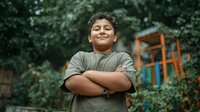 Young Hispanic boy with crossed arms smiling confidently outdoors in park playground setting with natural green foliage background for youth empowerment concepts.