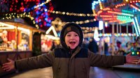 Young boy with arms outstretched enjoying festive carnival atmosphere with colorful lights and carousel in background during evening celebration.
