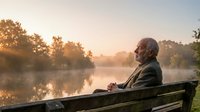 Senior Caucasian man sitting peacefully on wooden bench by misty lake at golden hour sunrise, enjoying tranquil morning nature scene for retirement lifestyle.