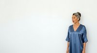 Mature woman with gray hair wearing blue satin shirt standing against white wall, looking away. Suitable for lifestyle, wellness, and professional content.