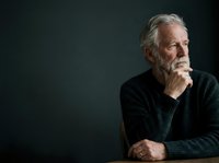 Senior man with gray hair and beard, wearing dark sweater, hand on chin in thoughtful pose against black background. Suitable for healthcare, wellness, and lifestyle editorial use.