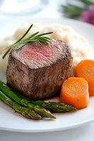 Juicy medium-rare beef tenderloin steak served with mashed potatoes, glazed carrots, grilled asparagus and fresh rosemary garnish on white plate.