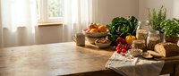 Rustic wooden kitchen table with fresh vegetables, herbs, bread and natural ingredients in warm sunlight for healthy cooking and meal preparation.